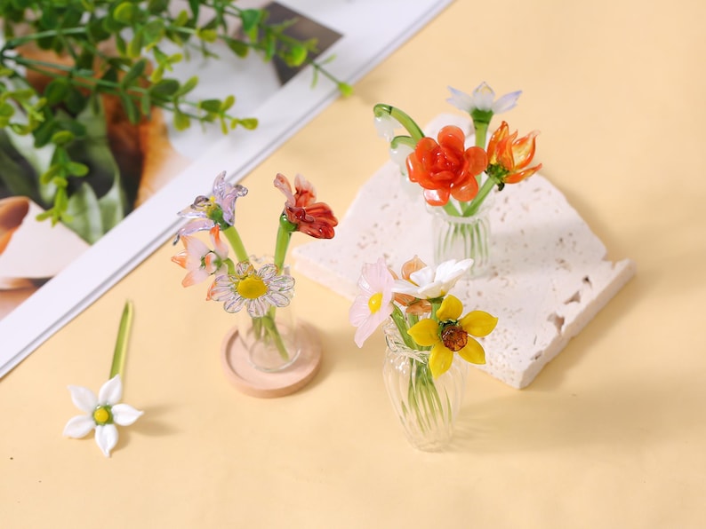 May include: Miniature glass flower bouquets in clear vases. The bouquets feature colourful flowers in shades of red, orange, yellow, pink, and white. One flower is separate. The vases sit on a beige surface.