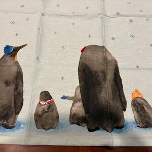 Puede incluir: Tela azul claro con una ilustraci&oacute;n de acuarela de ping&uuml;inos en una escena nevada. Los ping&uuml;inos llevan sombreros y bufandas de varios colores, incluyendo azul, rojo y naranja. Los copos de nieve est&aacute;n dispersos.