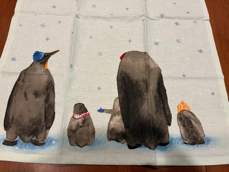 Puede incluir: Tela azul claro con una ilustraci&oacute;n de acuarela de ping&uuml;inos en una escena nevada. Los ping&uuml;inos llevan sombreros y bufandas de varios colores, incluyendo azul, rojo y naranja. Los copos de nieve est&aacute;n dispersos.