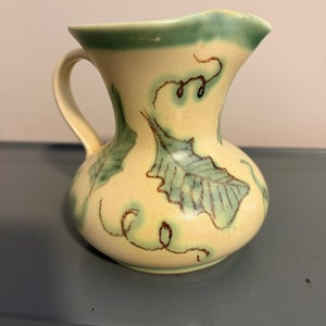 May include: A cream-colored ceramic pitcher featuring a hand-painted design of green leaves and brown swirling vine accents. The pitcher has a curved handle, a wide flared rim with a green painted edge, and a rounded body with a smooth finish.