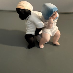 May include: A vintage porcelain figurine depicting two figures. One figure is wearing a white outfit and a cream-colored head covering. The other figure is wearing a white outfit and a blue head covering. The figurine is on a gray surface.
