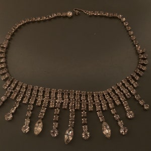 K&ouml;nnte beinhalten: Eine silberne Strasskette mit einem kaskadierenden Design. Die Kette hat eine Reihe von Strasssteinen auf der Vorderseite und eine Reihe von herabh&auml;ngenden Strasssteinen.