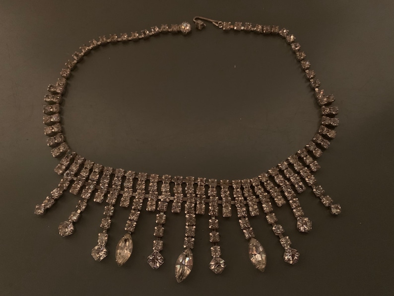 K&ouml;nnte beinhalten: Eine silberne Strasskette mit einem kaskadierenden Design. Die Kette hat eine Reihe von Strasssteinen auf der Vorderseite und eine Reihe von herabh&auml;ngenden Strasssteinen.