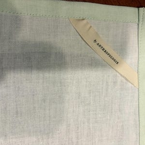 Puede incluir: Primer plano de una toalla de lino verde claro con un borde verde p&aacute;lido. Una etiqueta color crema con el texto "by ANTHROPOLOGIE" est&aacute; cosida en la esquina de la toalla.