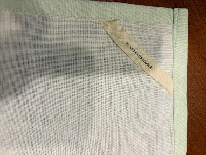 Puede incluir: Primer plano de una toalla de lino verde claro con un borde verde p&aacute;lido. Una etiqueta color crema con el texto "by ANTHROPOLOGIE" est&aacute; cosida en la esquina de la toalla.