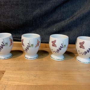 May include: Four white ceramic egg cups with a butterfly and grape design. Each cup has a rounded body, a pedestal base, and a decorative rim. The butterflies are in shades of orange and pink, and the grapes are purple.