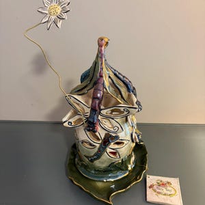 May include: A decorative ceramic candle holder with a dragonfly design. The holder is cream-colored with blue, green, and gold accents. A white flower on a wire extends from the top. The holder sits on a green leaf-shaped base.