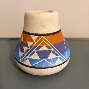May include: A small, rounded ceramic vase with a matte off-white finish. The body features horizontal bands of orange, purple, and blue, decorated with incised white geometric triangle patterns and thin black lines encircling the vessel.