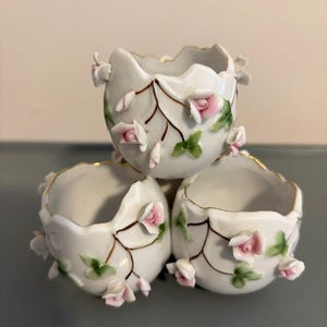 May include: Three white porcelain egg-shaped containers with pink rose accents and gold trim. The containers have a cracked eggshell design with pink roses and green leaves. The containers are stacked on top of each other.