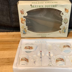 May include: A Beatrix Potter gardening set in a white box with a white background. The set includes a small teapot, a sugar bowl, a creamer, and four teacups. The teacups are decorated with Beatrix Potter characters. The box has a picture of Peter Rabbit on the front.