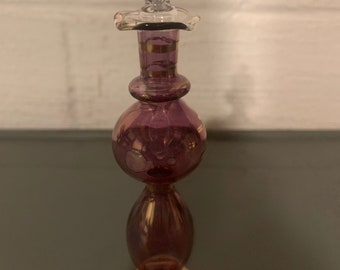 May include: A purple glass bottle with a gold rim and a clear glass stopper. The bottle has a round body and a long, slender neck.