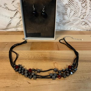 May include: A black necklace with three strands of small black beads and larger, dark gray, cube-shaped beads. The necklace has red beads interspersed throughout. The necklace is displayed on a wooden surface. A pair of black earrings are in a white box.