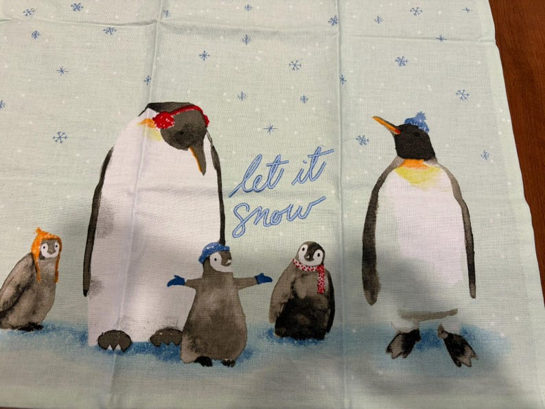 Puede incluir: Tela azul claro con una ilustraci&oacute;n de una familia de ping&uuml;inos. Los ping&uuml;inos llevan gorros y bufandas, con las palabras "Let it Snow" en escritura azul. Los copos de nieve est&aacute;n esparcidos por el fondo.
