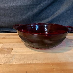 May include: A ruby red glass bowl with two handles. The bowl has a textured, ribbed design around the sides and a geometric pattern on the bottom. The bowl is sitting on a wooden surface.