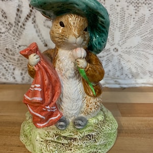 May include: A ceramic figurine of Peter Rabbit wearing a green hat and holding a red scarf and a white flower. The figurine is standing on a green base with brown accents.