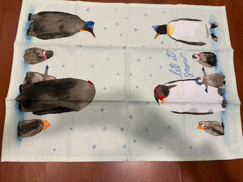 Puede incluir: Un pa&ntilde;o de cocina verde claro con un tema invernal. El pa&ntilde;o presenta ilustraciones de ping&uuml;inos con sombreros y orejeras, con las palabras "Let it snow" en escritura azul. Peque&ntilde;os copos de nieve azules est&aacute;n esparcidos por el pa&ntilde;o.