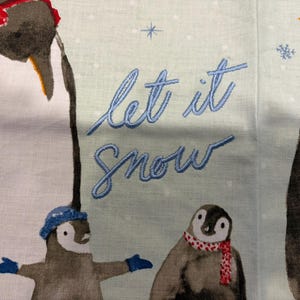Puede incluir: Tela azul claro con texto bordado &laquo;Let it Snow&raquo;. La tela presenta ilustraciones de ping&uuml;inos con sombreros y bufandas, con copos de nieve esparcidos por el fondo. Los ping&uuml;inos est&aacute;n en varias poses.