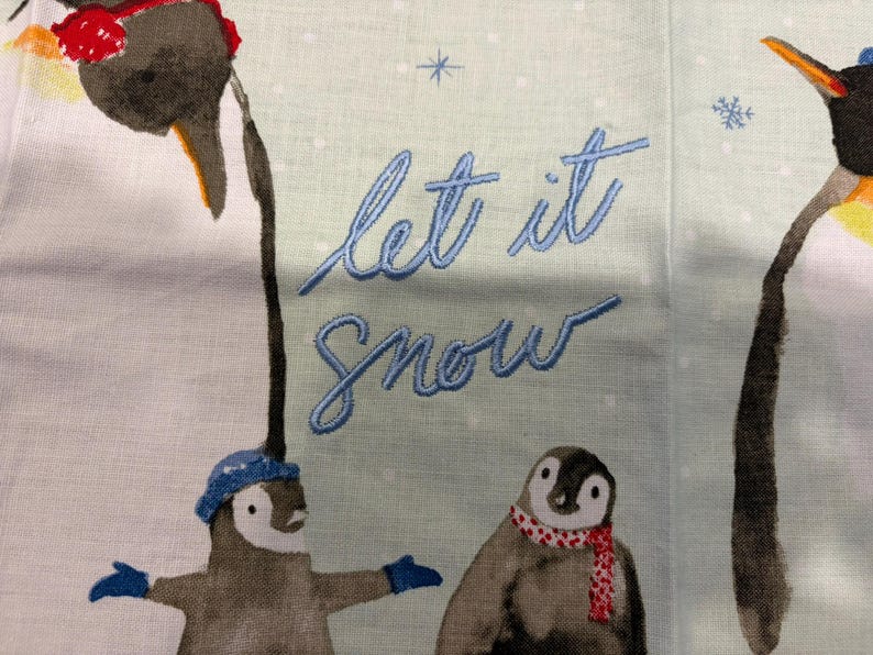Puede incluir: Tela azul claro con texto bordado &laquo;Let it Snow&raquo;. La tela presenta ilustraciones de ping&uuml;inos con sombreros y bufandas, con copos de nieve esparcidos por el fondo. Los ping&uuml;inos est&aacute;n en varias poses.