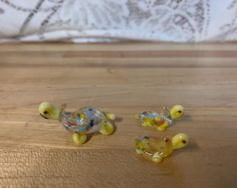 以下が含まれることがあります： 3つのガラス製の亀の置物で、それぞれサイズが異なります。一番大きな亀は黄色で、青と白の斑点があります。中くらいの亀は黄色で、赤と白の斑点があります。一番小さな亀は黄色で、黒と白の斑点があります。