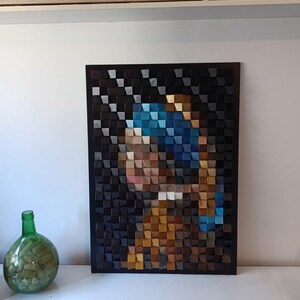 Peut inclure: Une œuvre d'art en mosaïque de bois représentant la célèbre peinture "La Jeune Fille à la Perle". L'image est créée à partir de petits blocs de bois carrés de différentes couleurs. L'œuvre est encadrée en noir et est exposée sur une surface blanche.