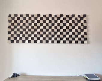 May include: A rectangular wall art piece composed of numerous small, individual wooden blocks arranged in a checkered pattern of alternating black and white squares, creating a three-dimensional, undulating surface texture.
