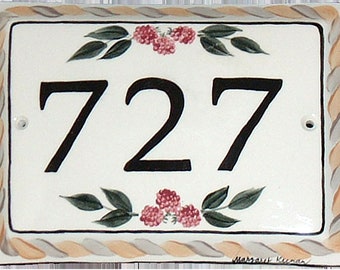 May include: White ceramic house number plaque with a black painted number "727" and a decorative border of a braided rope design in beige and gray. The top and bottom of the plaque are decorated with green leaves and pink berries.