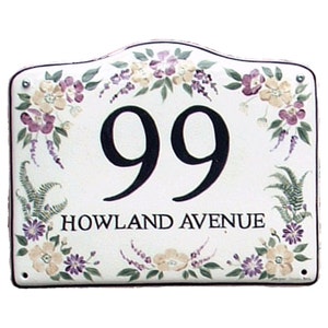 Peut inclure: Panneau de numéro de maison en céramique blanche avec un motif floral dans les tons de violet, vert et jaune. Le numéro 99 est peint en noir et le texte "HOWLAND AVENUE" est écrit en noir sous le numéro.