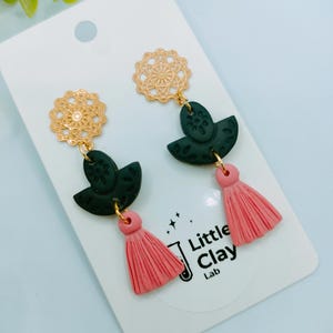 May include: Dangle earrings with gold filigree tops, dark green leaf shapes, and pink tassel drops. The earrings are presented on a white card with the text "Little Clay Lab".