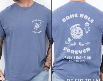 May include: A blue jean colored t-shirt featuring a white graphic of a cartoon golf ball wearing a cap, holding a golf club and a beer glass. The text reads SAME HOLE FOREVER, JASON'S BACHELOR, and NASHVILLE, 2026. The design appears on the front chest and back.