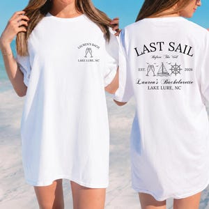 May include: A white t-shirt featuring black text and graphics. The front chest displays "LAUREN'S BACH" above two champagne flutes and "LAKE LURE, NC". The back features "LAST SAIL Before The Veil", a sailboat, ship wheel, and "Lauren's Bachelorette".