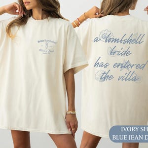 May include: An ivory t-shirt featuring blue text and graphics. The front displays "Bride Bombshell" with a cocktail icon and "Olivia's Bach Austin, TX". The back features the text "a bombshell bride has entered the villa" with various seashells.