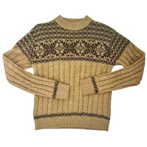 May include: Tan sweater with a mock neck and long sleeves. The sweater features a cable knit design on the body and striped pattern on the sleeves. A dark brown snowflake pattern is on the upper chest and shoulders.
