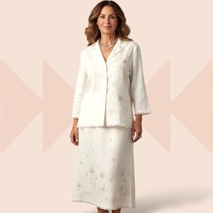 May include: Off-white linen skirt suit with floral embroidery. The suit includes a long-sleeved jacket with a notched collar and a matching knee-length skirt. The jacket and skirt have a fringed hem. The floral embroidery is in a neutral color.