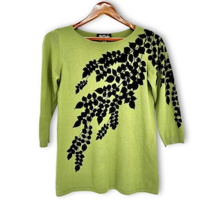 May include: A pale green sweater with three-quarter length sleeves. The sweater has a black leaf pattern across the front and right shoulder. It is displayed on a wooden hanger.