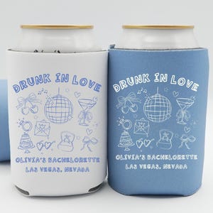 May include: White and blue can coolers with the text "DRUNK IN LOVE" and various illustrations. The text "Olivia's Bachelorette Las Vegas, Nevada" is also printed on the coolers. One cooler is white, the other is blue.