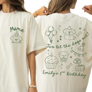 May include: Ivory-colored t-shirt with a moss green design. The front features the word "Mama" above a dog illustration with a cupcake. The back has a dog-themed birthday design with text that reads "Two let the dogs out?" and "Emily's 1st Birthday".