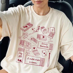 May include: Ivory-colored t-shirt with a burgundy design featuring matchboxes, hearts, and text. The text includes "July 2025", "Emma's Bachelorette", and "A match made in heaven".
