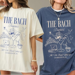 May include: Ivory and denim t-shirts with a blue graphic that reads "THE BACH Diaries" above a martini glass with a woman inside. The text "Me? The Bride? Shut up! CHARLESTON, SC" is below the graphic.