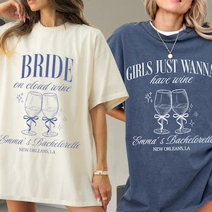 May include: Two t-shirts, one ivory and one denim, with wine-themed graphics. The ivory shirt reads "BRIDE on cloud wine" with wine glass illustrations. The denim shirt says "GIRLS JUST WANNA have wine" with similar wine glass art. Both shirts include "Emma's Bachelorette, New Orleans, LA".