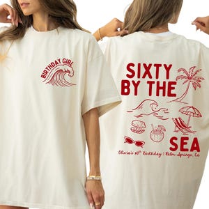 May include: An ivory t-shirt featuring a burgundy graphic design. The front displays a wave and the text "BIRTHDAY GIRL". The back features the text "SIXTY BY THE SEA", "Olivia's 60th Birthday | Palm Springs, Ca", and illustrations of a palm tree, wave, seashell, coconut drink, beach chair, umbrella, and sunglasses.