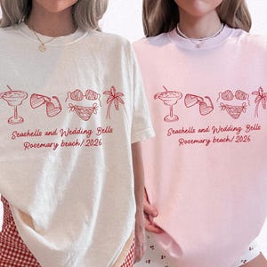 May include: Two t-shirts, one ivory and one pink, with a red graphic. The graphic includes a cocktail, sunglasses, a bikini, and a palm tree. Text reads "Seashells and Wedding Bells Rosemary beach/2026".