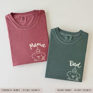 May include: Two t-shirts, one crimson and one moss green, each with a white graphic of a dog with a cupcake on its head. The crimson shirt says "Mama", and the moss green shirt says "Dad". Both shirts are Comfort Colors brand.