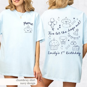 May include: Light blue chambray shirt with navy design. The front features a cartoon dog with the word "Mama". The back has a birthday theme with the text "Two let the dogs out?" and "Emily's 2nd Birthday".