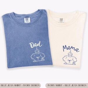 May include: Two Comfort Colors t-shirts, one blue jean and one ivory. The blue shirt has "Dad" and a dog graphic with a cupcake. The ivory shirt has "Mama" and a matching dog graphic. Both shirts are folded.