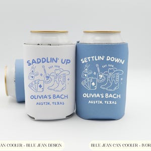 Op de afbeelding: Twee blikjeskoelers, &eacute;&eacute;n wit en &eacute;&eacute;n blauw, met een countrythema. De witte koeler heeft een blauw ontwerp met de tekst "Saddlin' Up", de andere heeft een wit ontwerp met de tekst "Settlin' Down". Beide zeggen "Olivia's Bach, Austin, Texas".