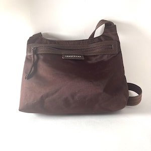 Puede incluir: Un bolso de hombro marrón con cremallera en la parte superior y una correa para el hombro. El bolso tiene forma rectangular con la base redondeada. El nombre de la marca "LONGCHAMP" es visible en la parte delantera.