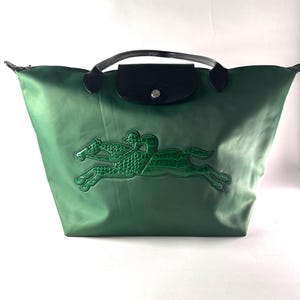 May include: Green tote bag with a black leather flap and handles. The bag features a textured green applique of a horse and rider. The bag is made of a shiny, durable material.