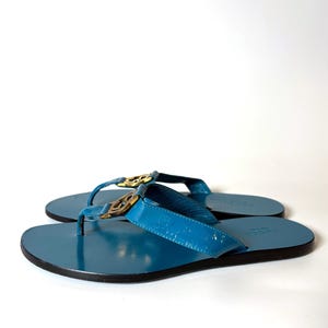 Side profile of the blue patent leather flat sandals with gold hardware on a white background.