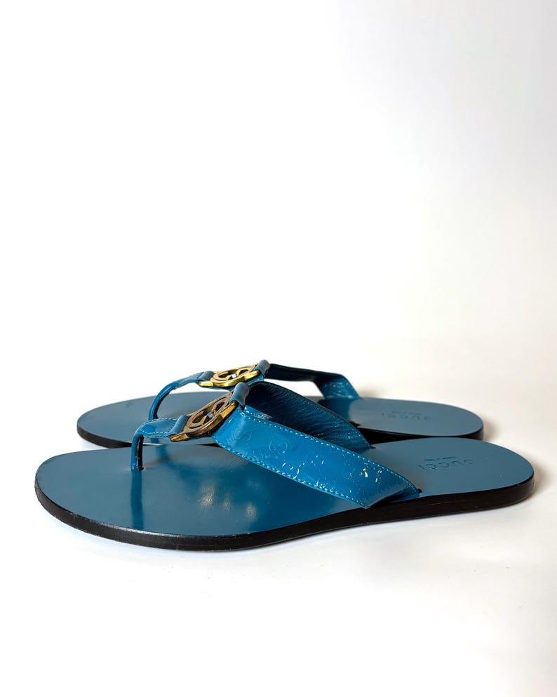 Side profile of the blue patent leather flat sandals with gold hardware on a white background.