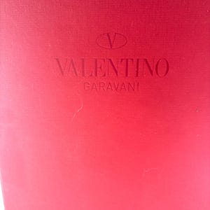 May include: A red box with the Valentino Garavani logo embossed on the front. The logo features a stylized 'V' above the brand name.